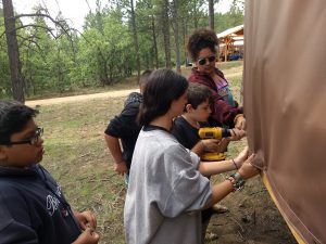 a few campers and a counselor work on tightening screws outside a yurt, using a drill