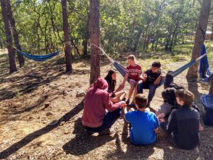 Small group of campers and staff, most sitting on the ground in the shade, two in a hammock