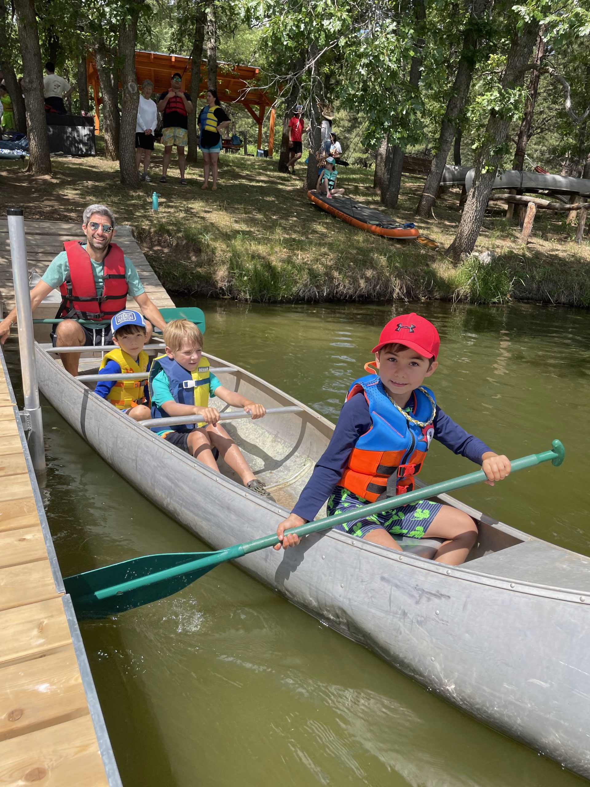 A father with three children in a canoe, oldest child has a paddle and is about to push off from the dock. Water front, trees, other campers, picnic structure in background. 