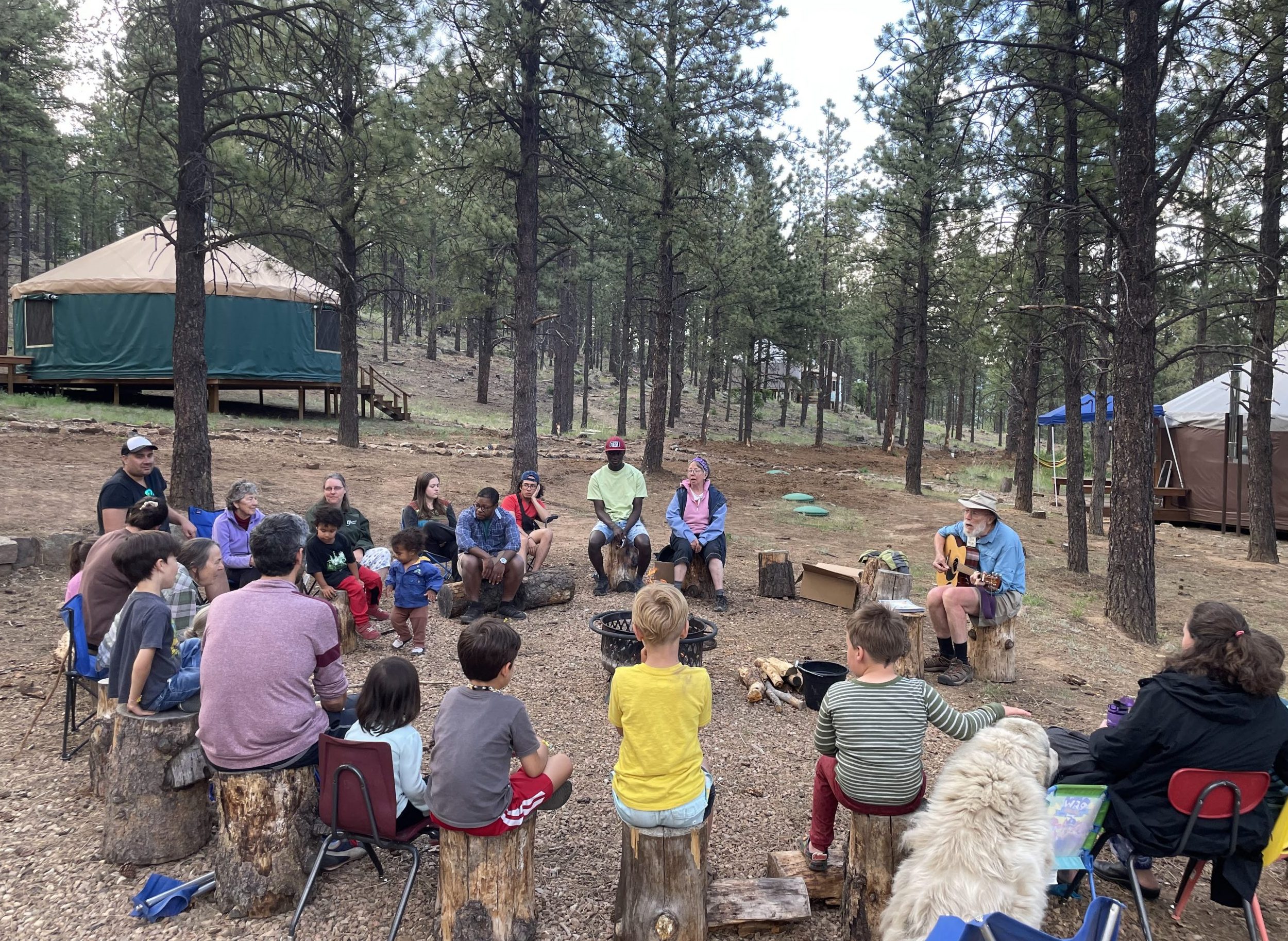 Group of families and staff sitting around the campfire, one with guitar, plus two ranch dogs. Green yurt and pine trees in background.