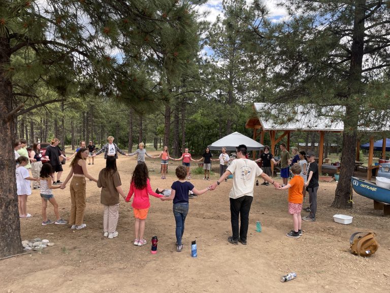 Circle of campers and staff holding hand, with pine trees and yurt in frame